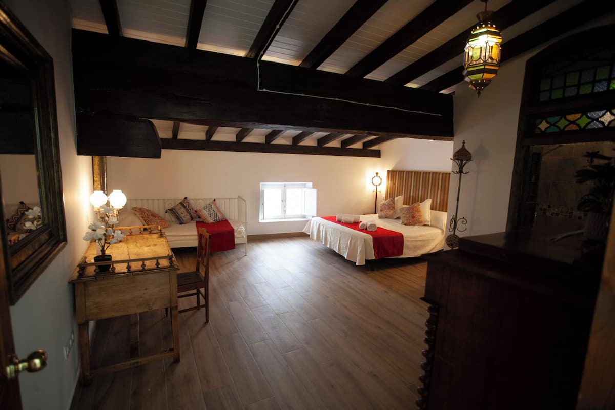A room with a wooden floor, a bed with a red bedspread, a bedside table with a lamp, and a window with daylight.