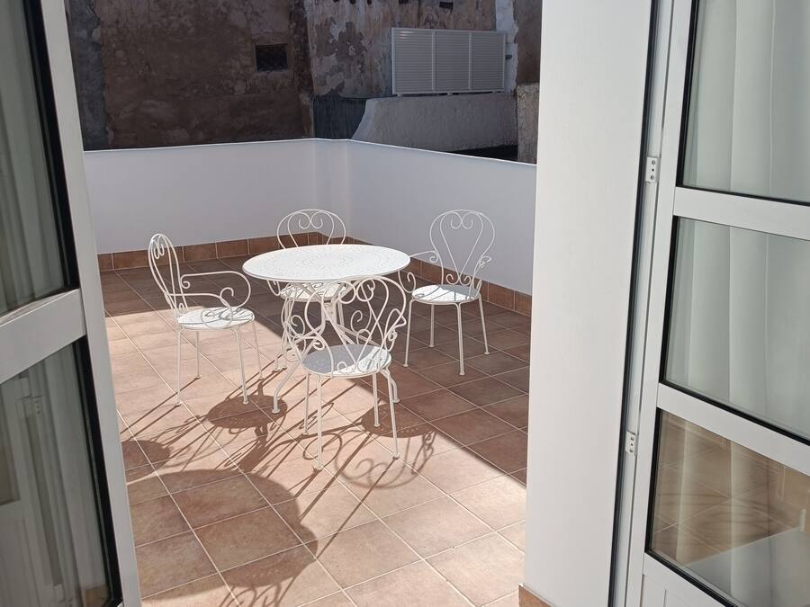 Un balcón abierto con una mesa y sillas de metal blanco, suelo de baldosas y vistas a edificios con cúpulas y antenas al fondo.