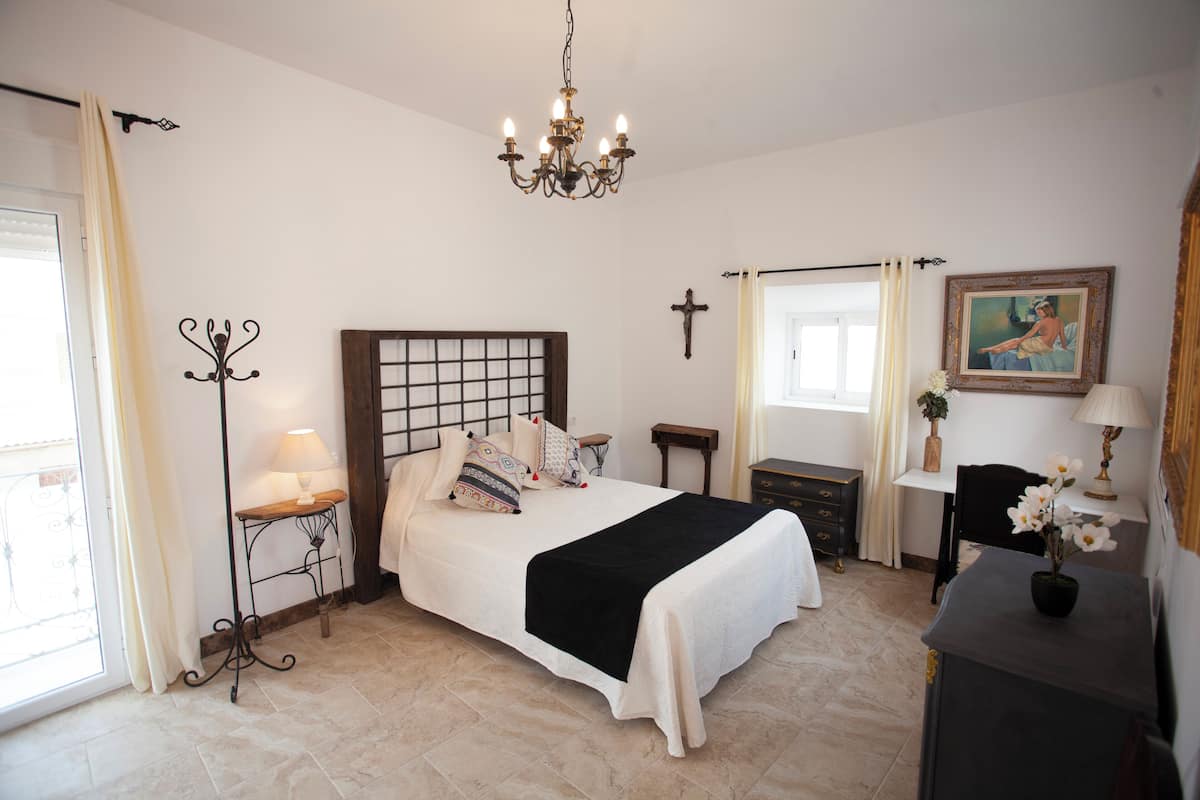 A bedroom with a bed, bedside tables, a desk, a chair, a cross on the wall, and a chandelier.
