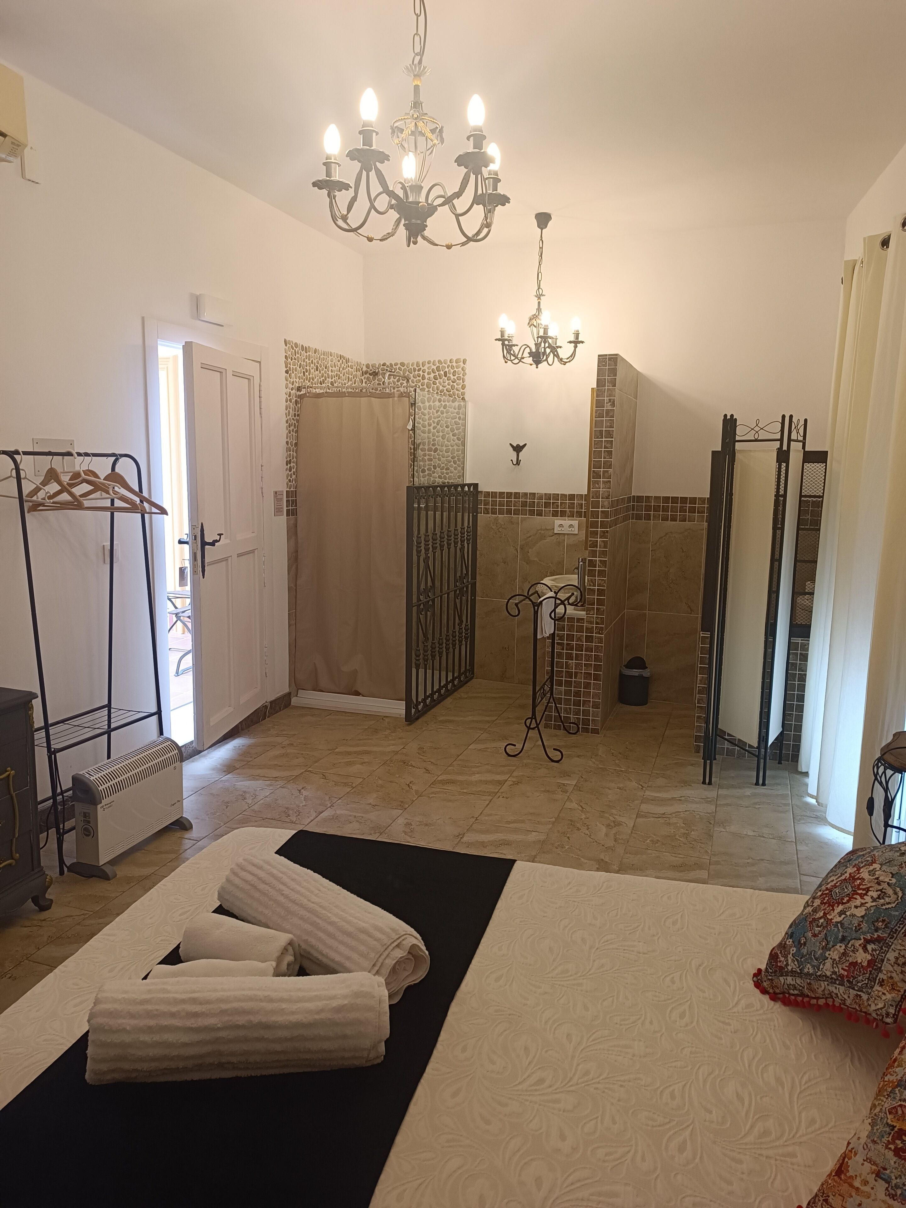 Une chambre d’hôtel comprenant un lit, un radiateur, un lustre, un espace douche et une salle de bain avec un mur carrelé.