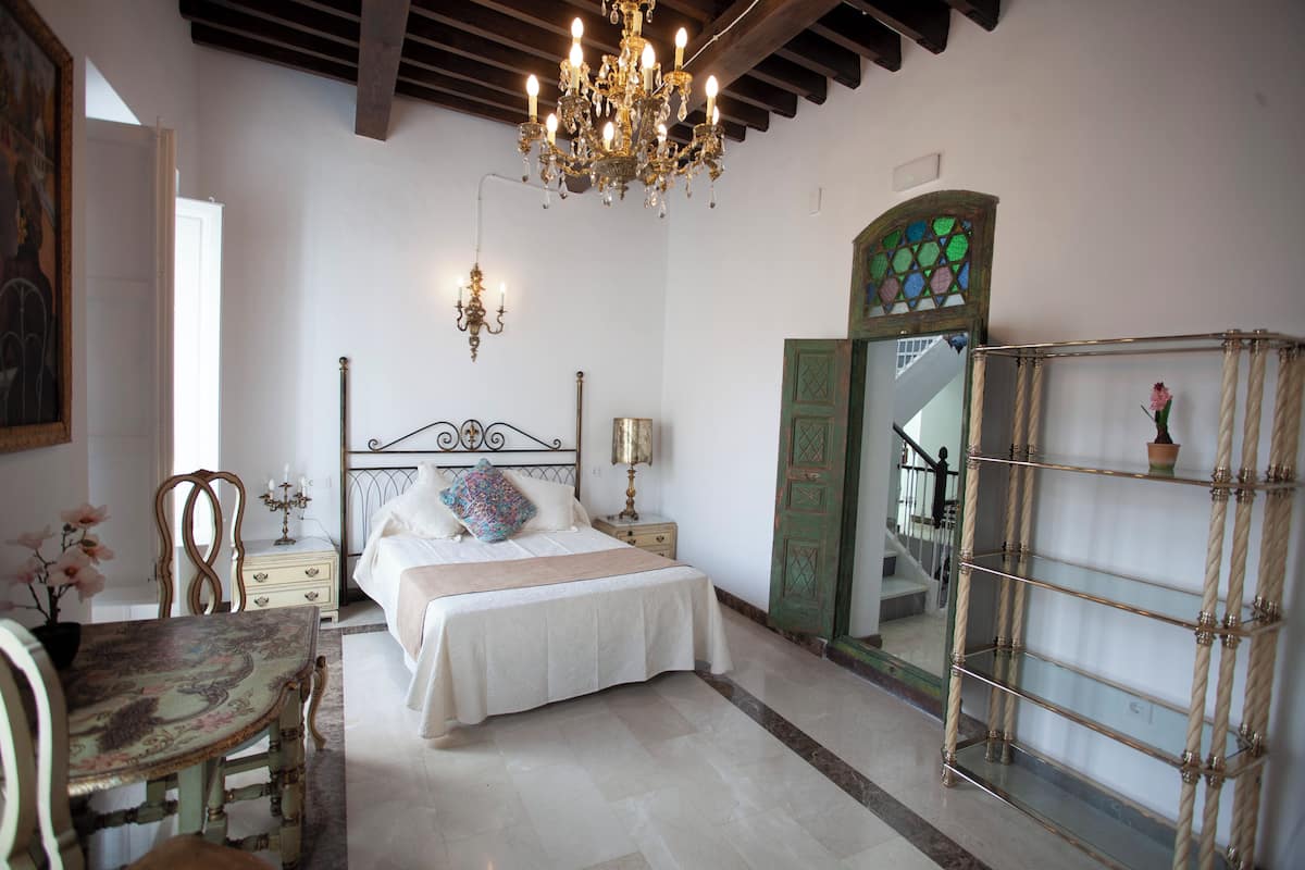 A bedroom with a bed, bedside tables, a chandelier, a mirror, and a chair.
