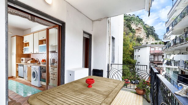 Un balcon avec une table en bois et un objet décoratif rouge, une cuisine équipée de machines à laver et une vue sur une région montagneuse.
