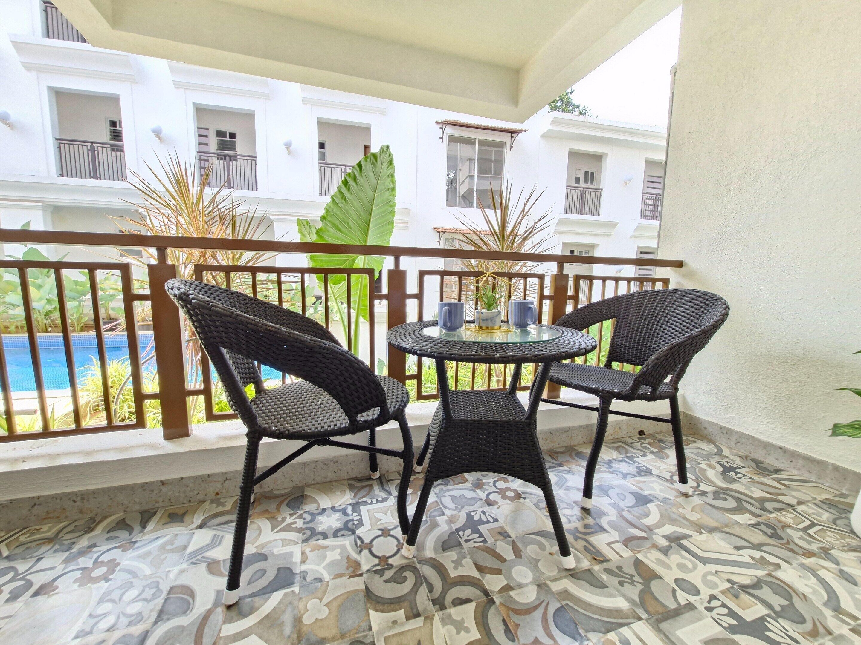 Un balcón con sillas de mimbre y una mesa de vidrio, con vistas a una piscina y un edificio con balcones.