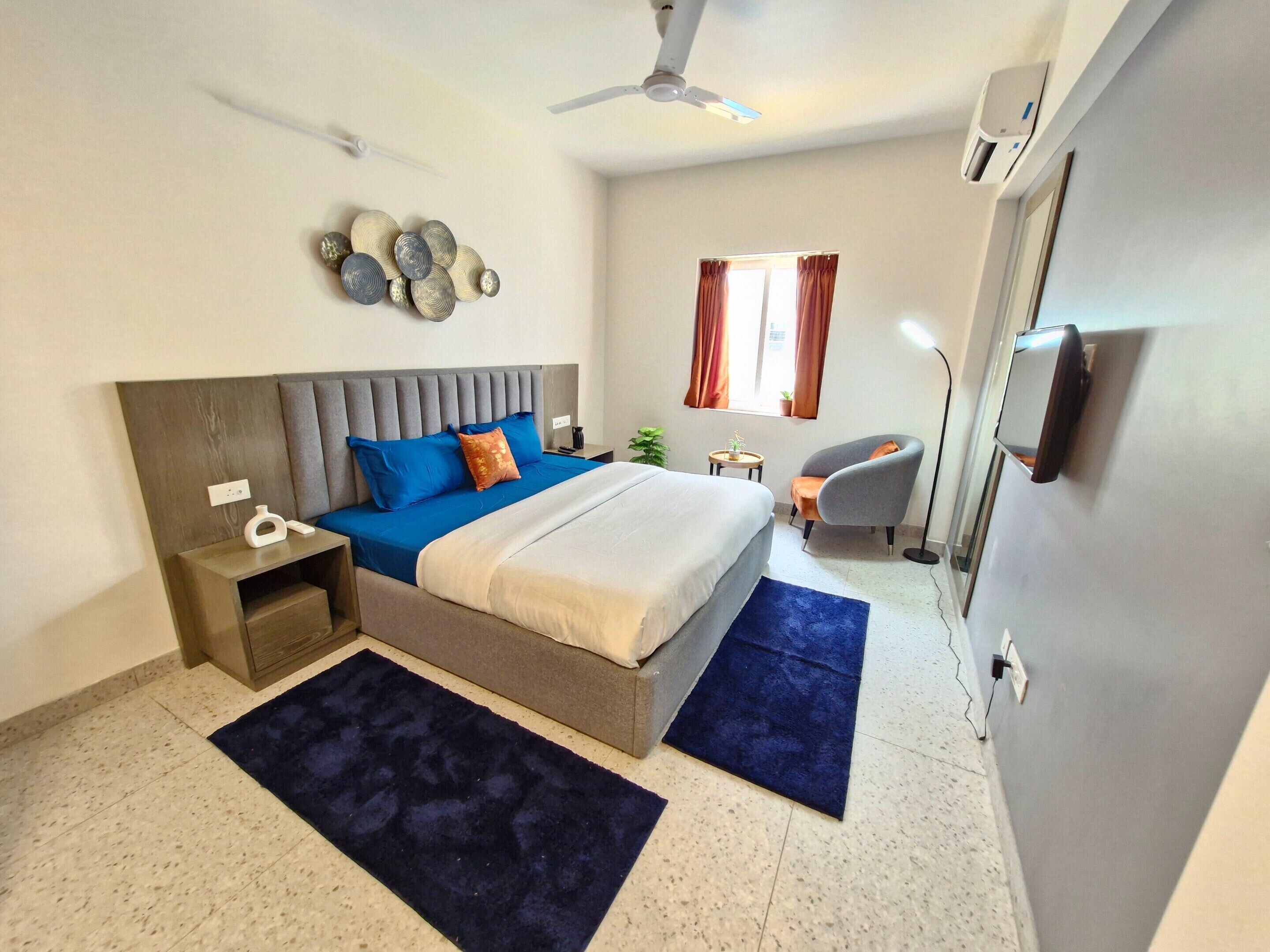 A modern bedroom with a large bed, a bedside table, a chair, a small table, a plant, and a window with curtains.