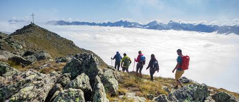 A group of hikers on a rocky mountain path with a cross on a hilltop, surrounded by clouds and distant mountains.