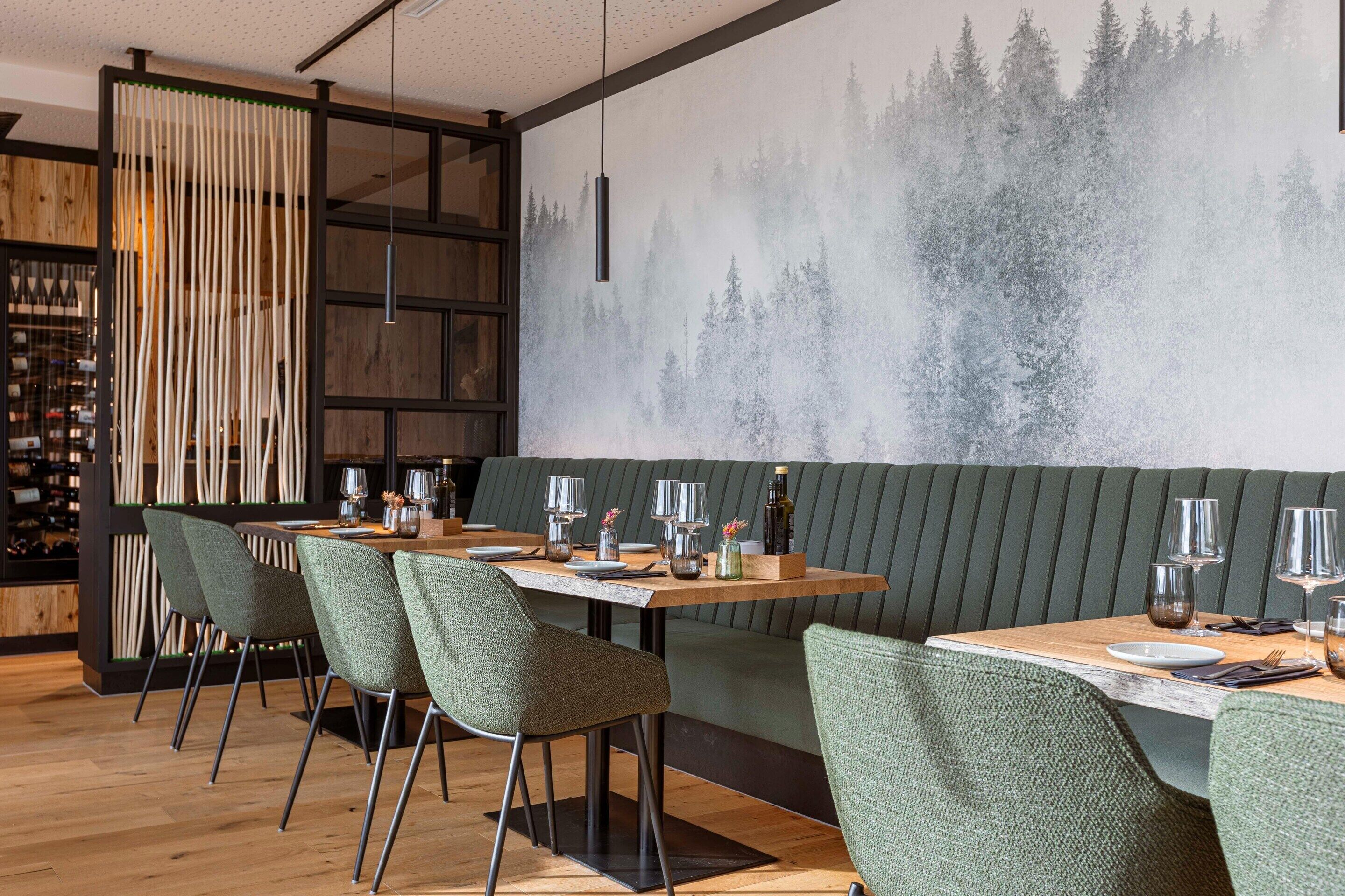Un restaurant avec une longue table en bois, des chaises à l’assise rembourrée de tissu vert et une fresque murale représentant une forêt.