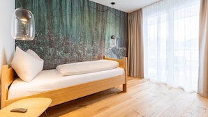 Ein modernes Zimmer mit einem hölzernen Bett, einem kleinen Tisch, einer Wandlampe und einem großen Fenster mit durchsichtigen Vorhängen.