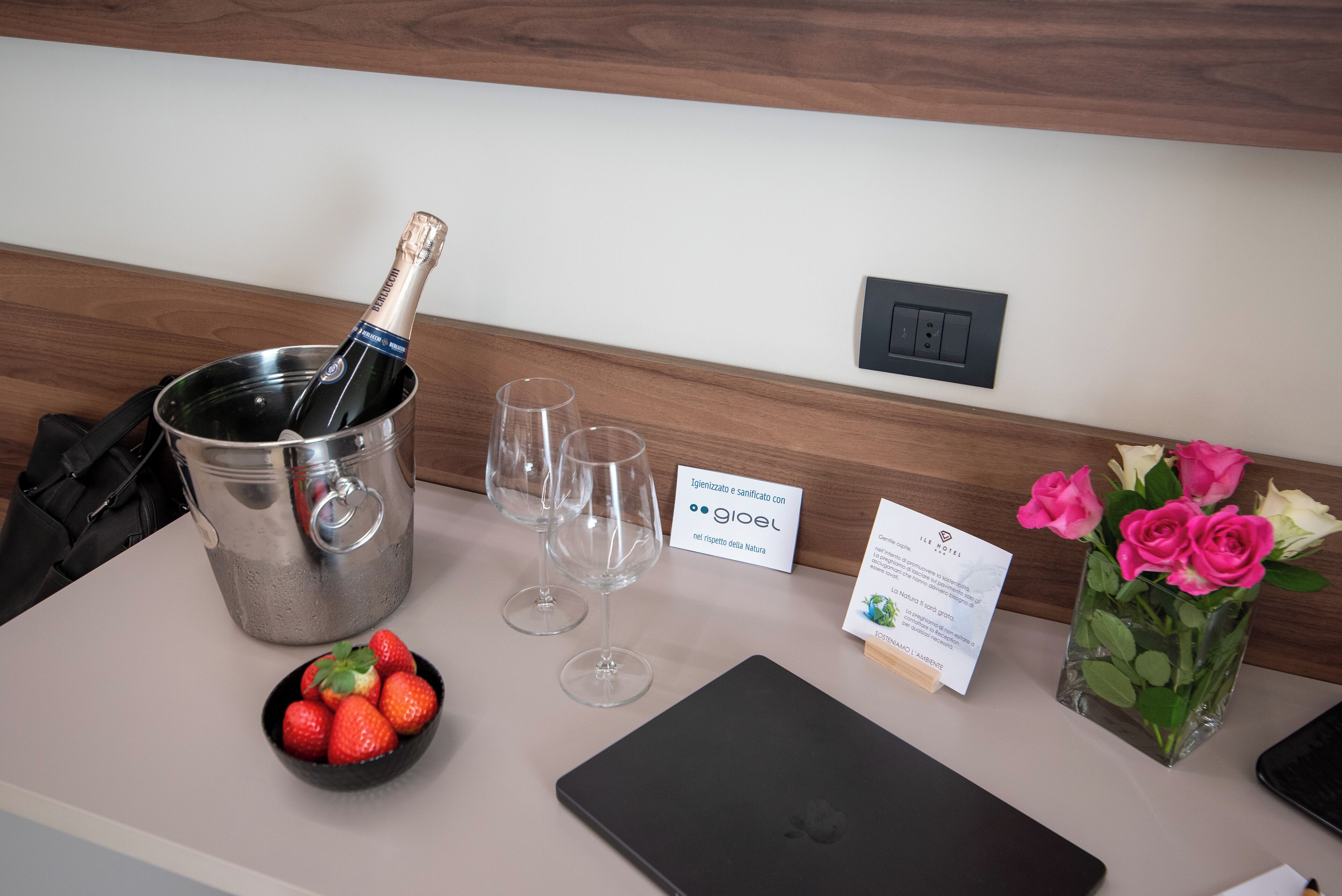 A hotel reception area with a champagne bottle in a bucket, two empty wine glasses, a bowl of strawberries, a laptop, a card, and a vase with flowers.