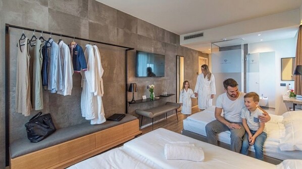 A hotel room with a large bed, a seating area with a bench, a TV mounted on the wall, and a clothing rack with various garments. - Ile Hotel (Peschiera del Garda)