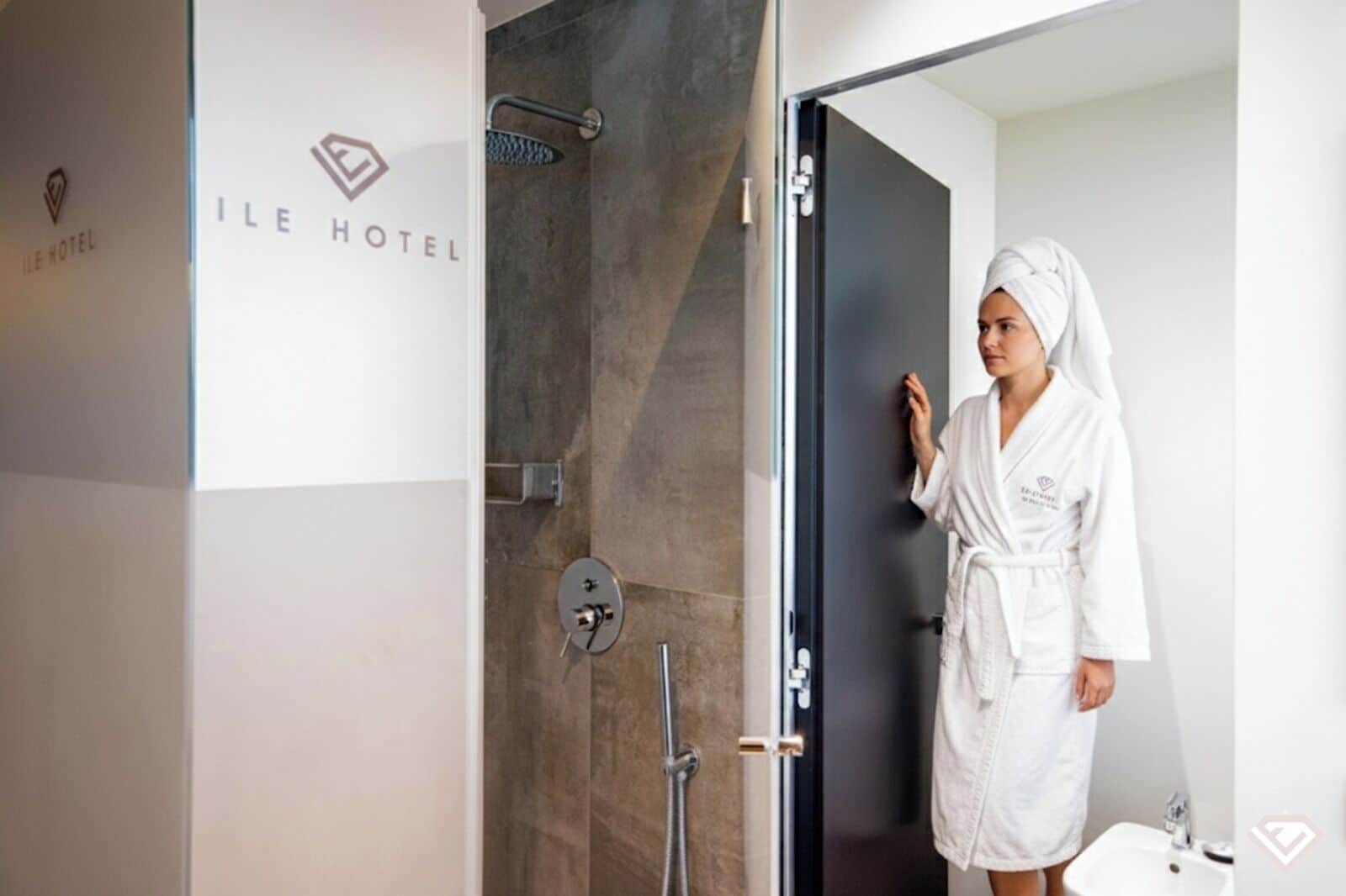 A person in a white robe standing in a modern bathroom with a shower and a sink.