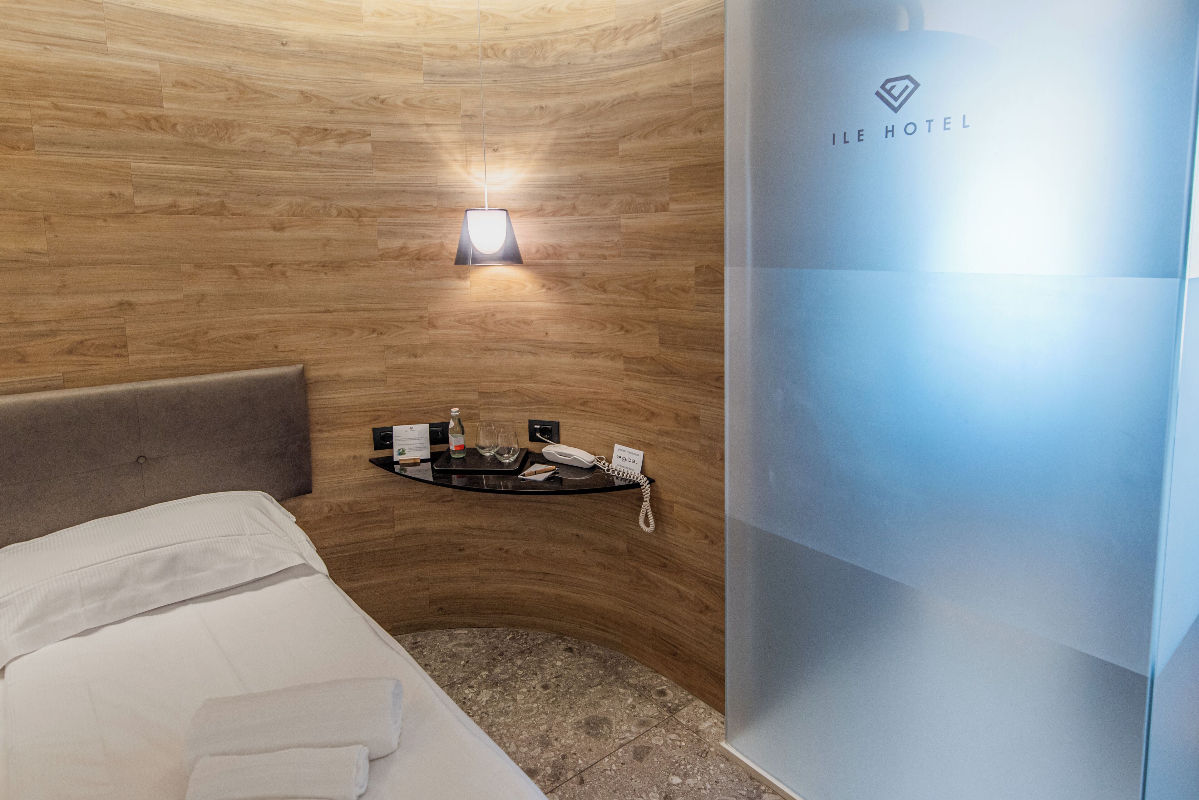 A hotel room with a wooden wall, a bed with white linens, a bedside table with a phone and a bottle, and a wall-mounted light.