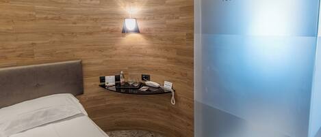A hotel room with a wooden wall, a bed with white linens, a bedside table with a phone and a bottle, and a wall-mounted light.