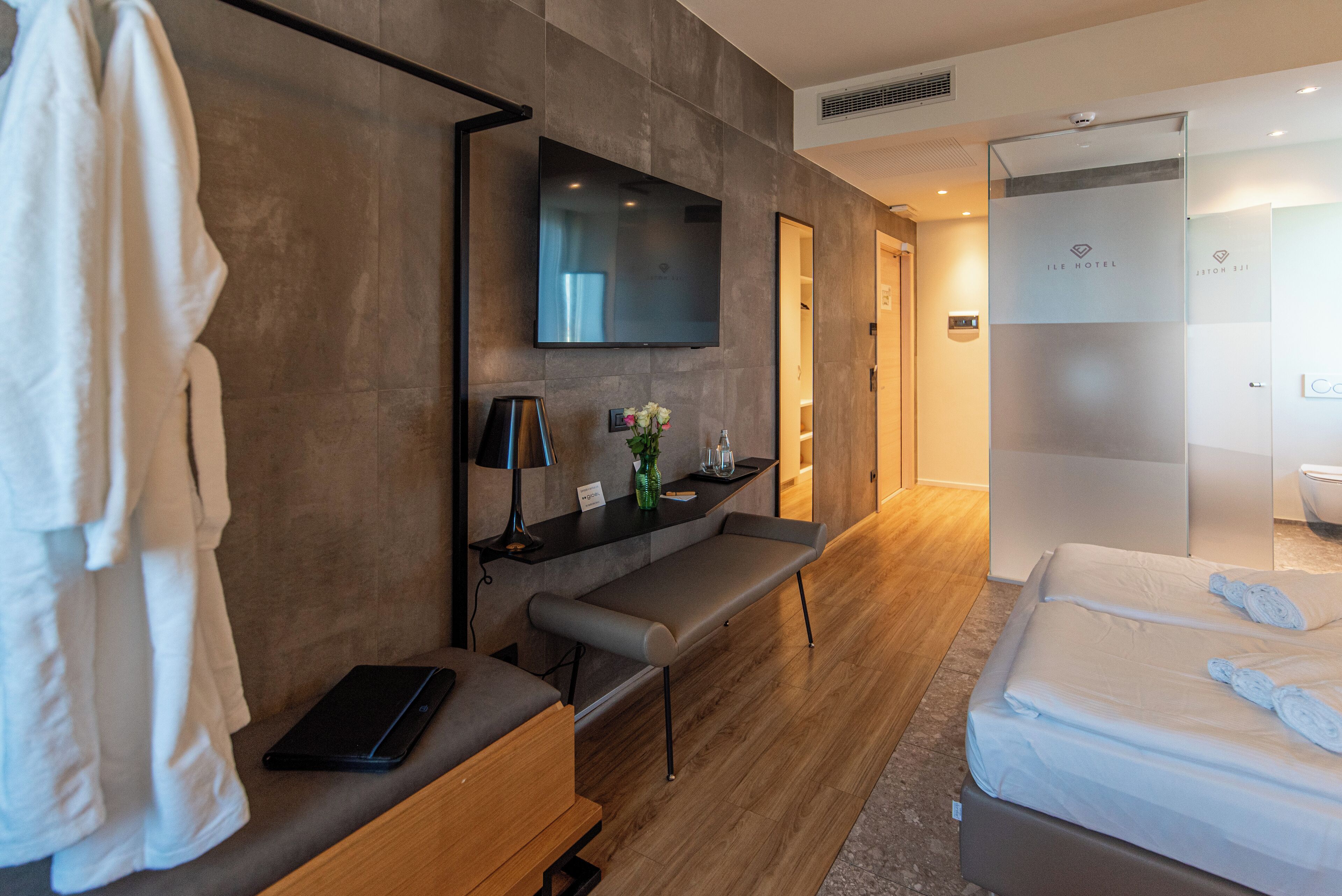 Una habitación de hotel moderna con una televisión de pantalla plana montada en la pared, un banco, una mesita de noche con lámpara y una cama con toallas.