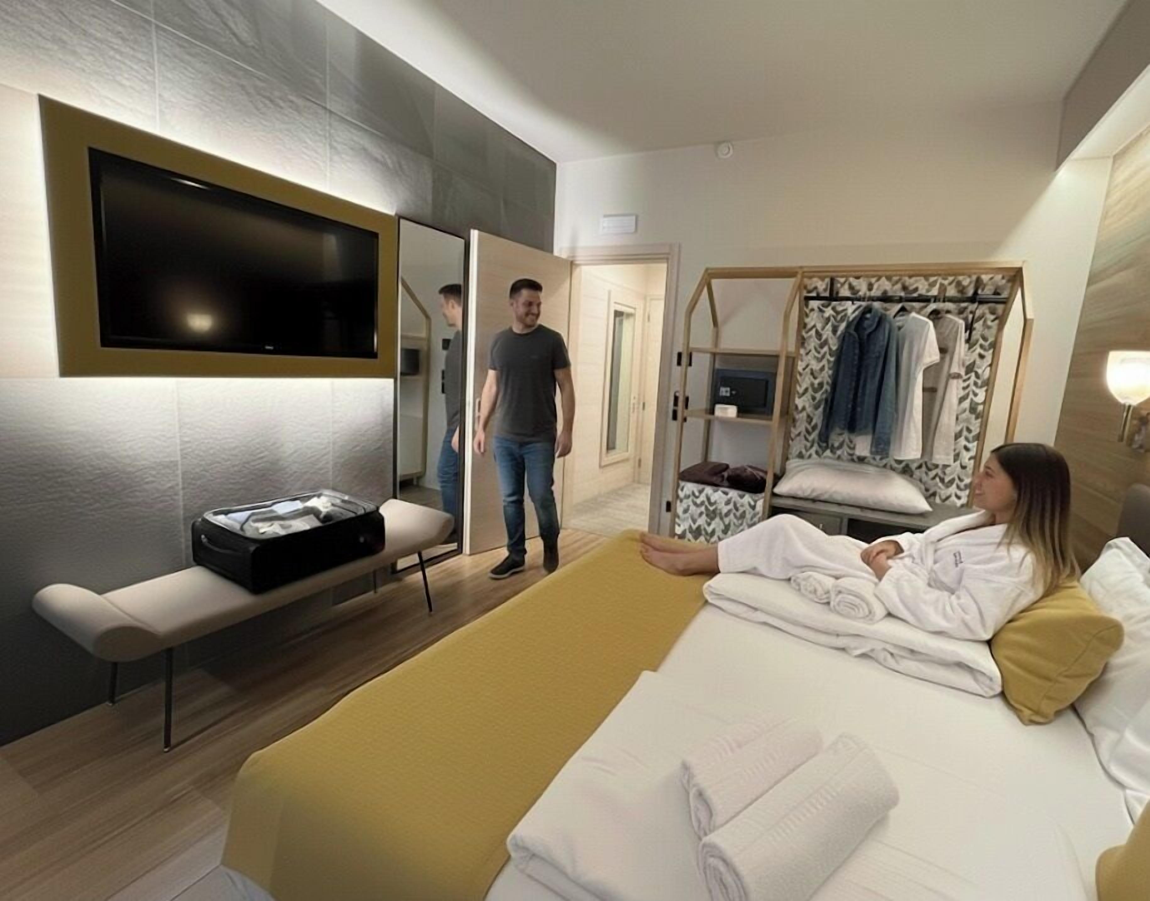 A modern hotel room with a bed, a TV, a bench, a mirror, and a wardrobe.