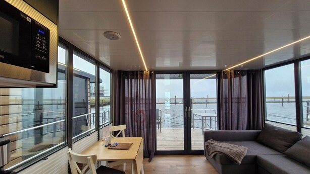 A modern interior with a dining area, sofa, and large windows offering a view of the water. - Houseboat Fjord del Sol with bio sauna and roof terrace in Wendtorf / Kiel Fjord (Wendtorf)