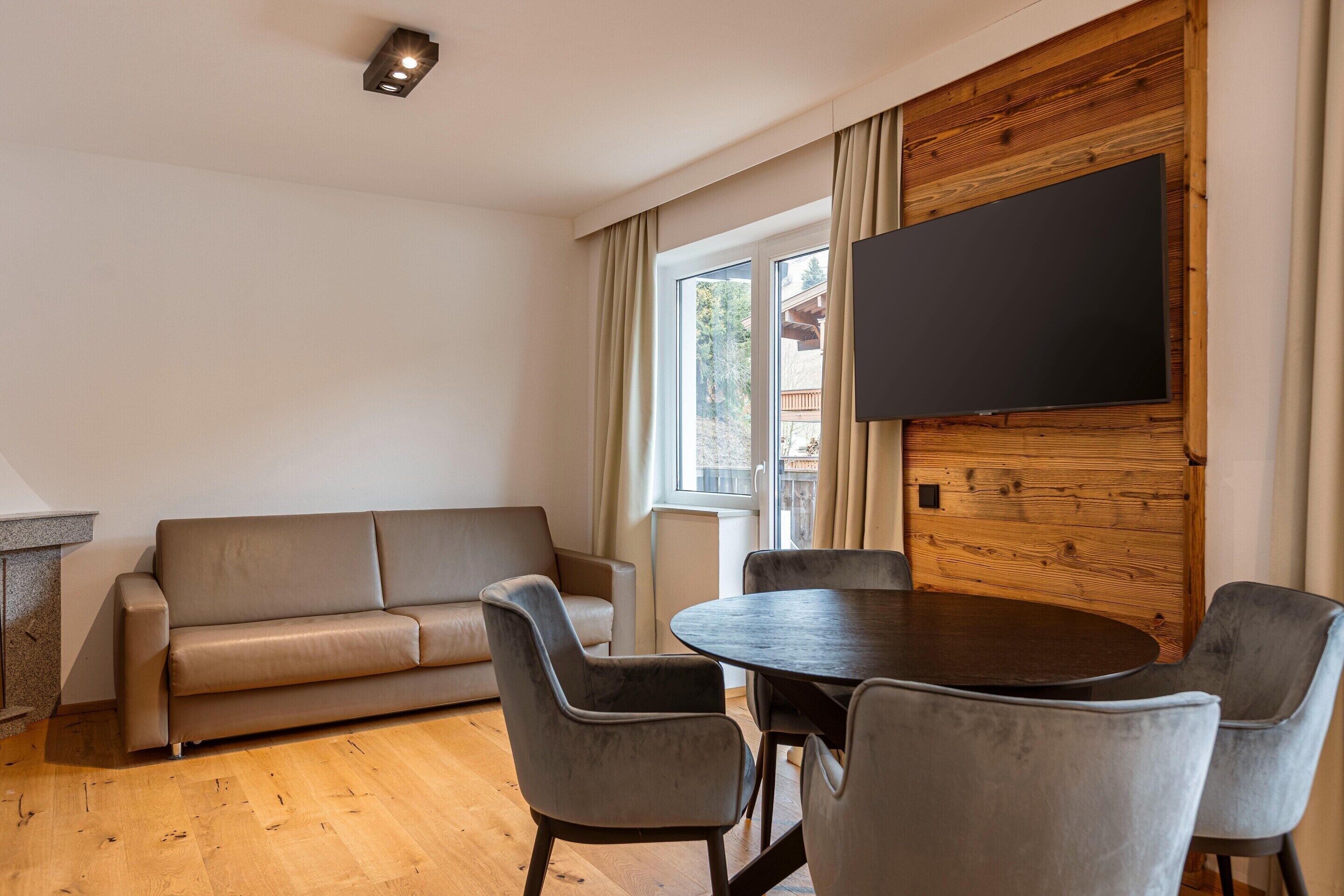 Una sala de estar moderna con un sofá, una mesa redonda con sillas, un televisor de pantalla plana montado en una pared de madera y una ventana con cortinas.