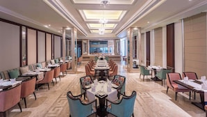 A spacious hotel dining area with multiple tables and chairs, a central chandelier, and a view of the kitchen in the background. - Park Plaza Jalandhar (Jalandhar)