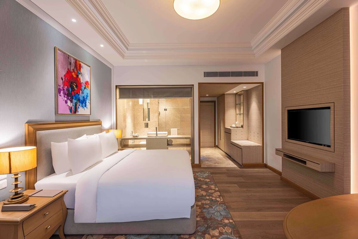 A modern hotel room with a large bed, a desk, a television, and a bathroom visible through an open door.