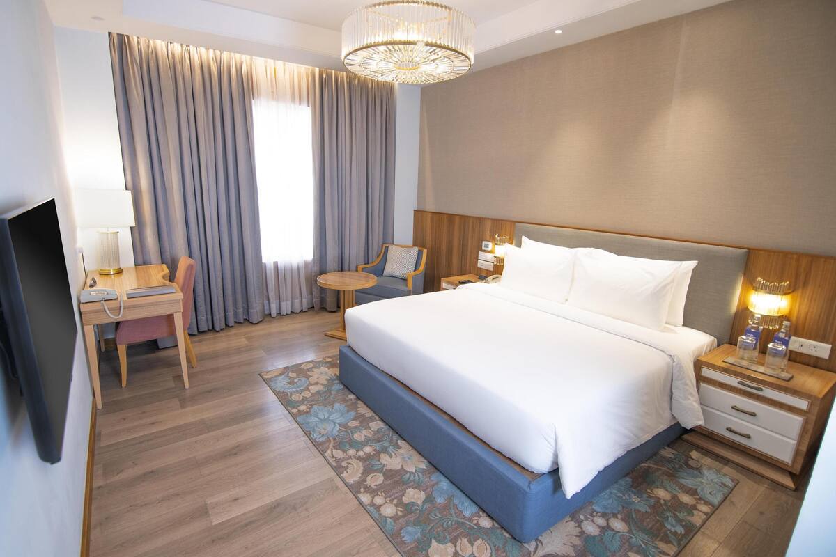 A hotel room with a large bed, a desk with a lamp, a chair, a bedside table with a lamp, and a TV.