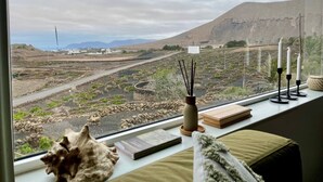A room with a view of terraced fields and mountains, a window with a decorative shell, a candle holder, and a book on the windowsill. - Casa MaVi - Vineyard with ocean view  (pool, privacy, scenic views, BBQ, AC)  (Tinajo)