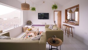 A modern living room with a sofa, coffee table, and a TV mounted on the wall. There are hanging plants and a large window providing a view of the city. - Casa MaVi - Vineyard with ocean view  (pool, privacy, scenic views, BBQ, AC)  (Tinajo)