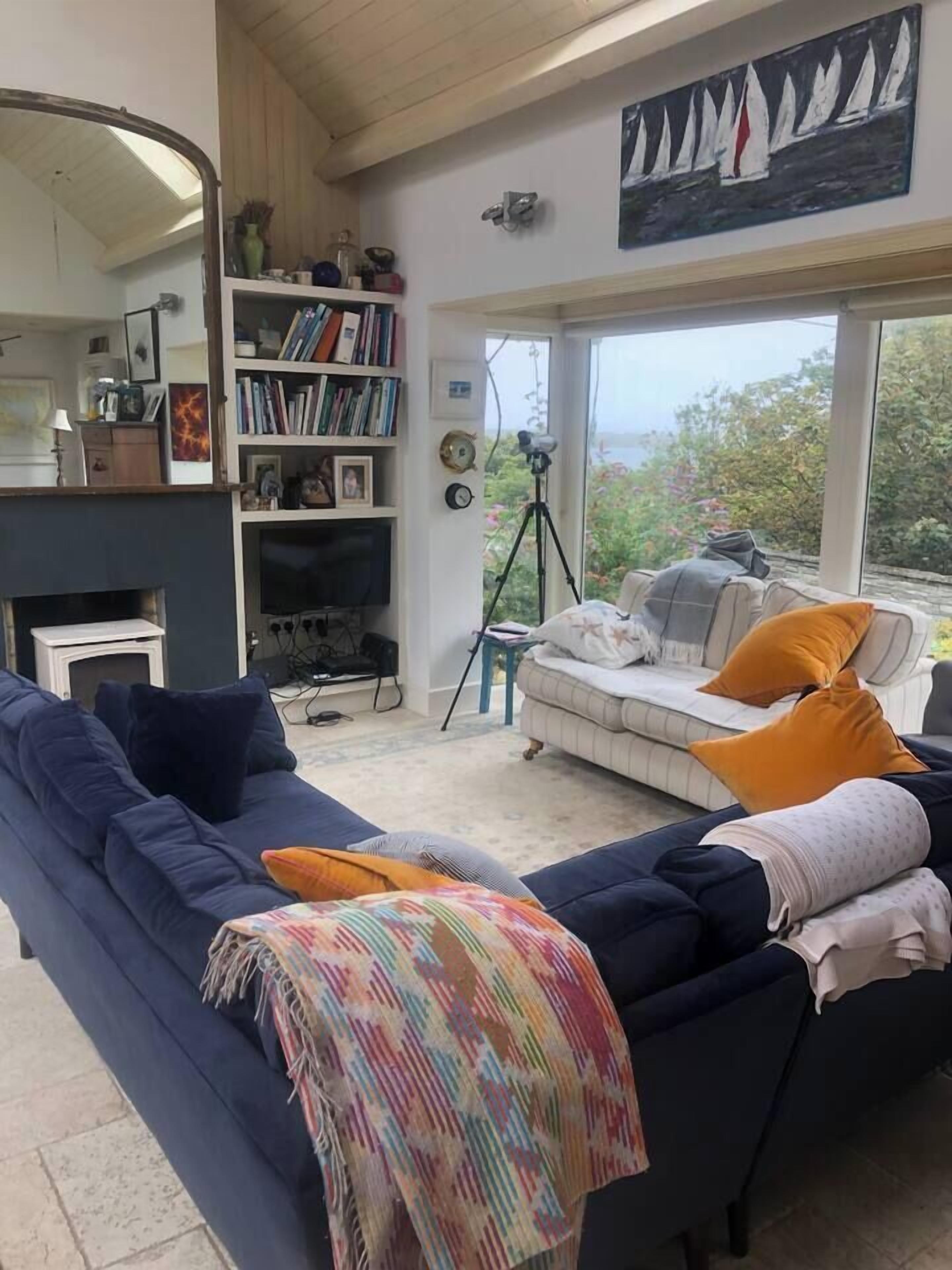 A living room with a blue sofa, a white sofa with orange cushions, a bookshelf, a fireplace, and a large window with a view of greenery.