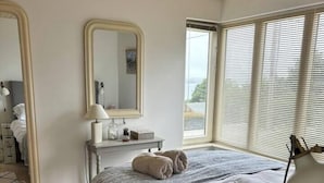 Ein Schlafzimmer mit einem großen Bett, einem Nachttisch mit Lampe, einem Spiegel und einem Fenster mit Jalousien.