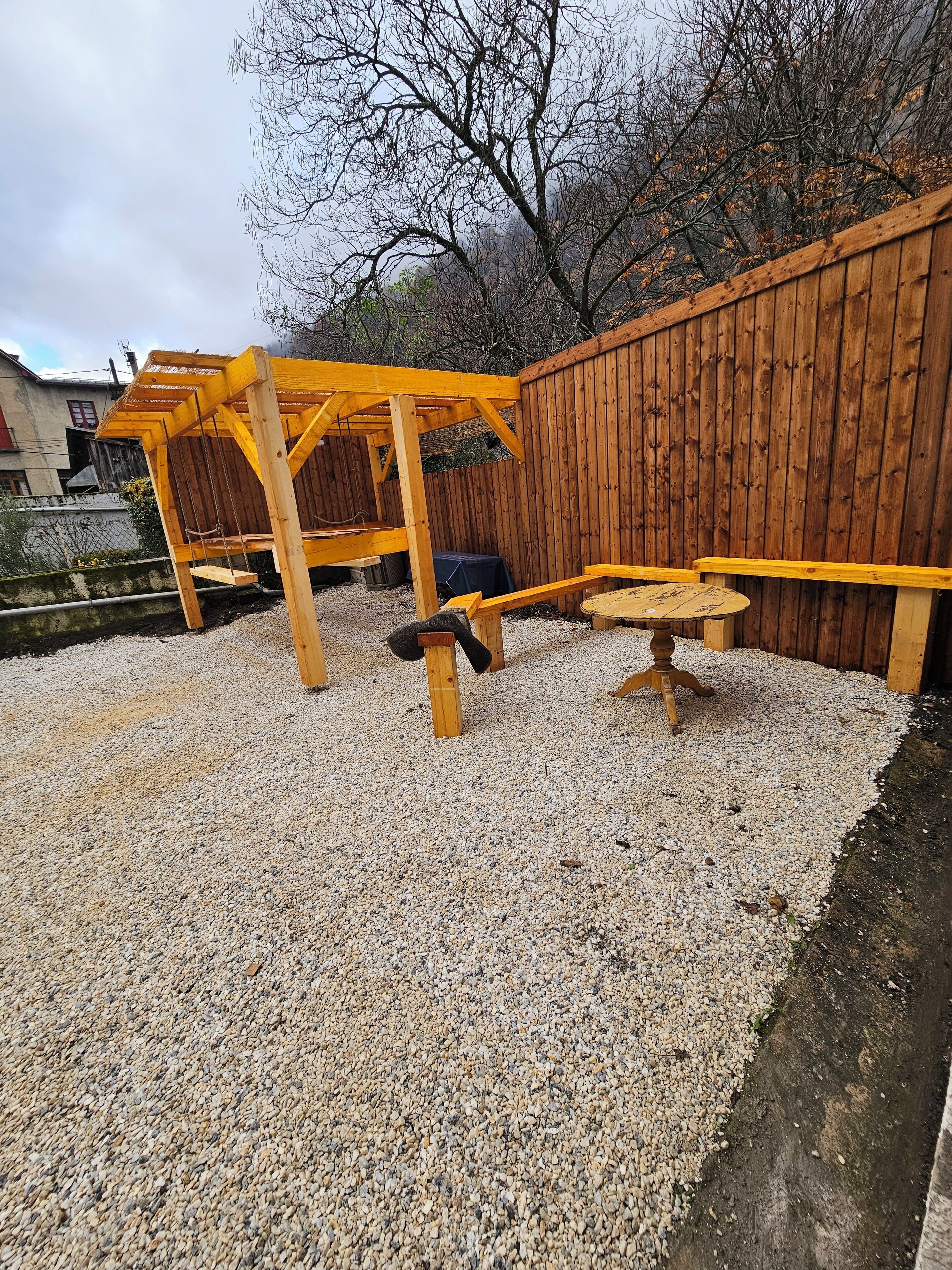 A wooden swing set and a bench in a gravel area with a wooden fence and trees in the background.