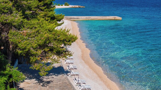 A beach with clear blue water, a stone pathway, and lounge chairs along the shore. - Hotel Waterman Kaktus (Supetar)