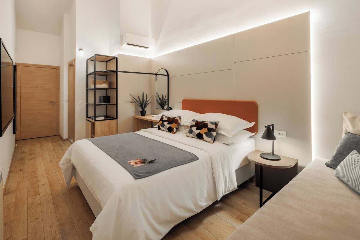 A modern bedroom with a wooden floor, a bed with white bedding, a nightstand with a lamp, and a small shelving unit.