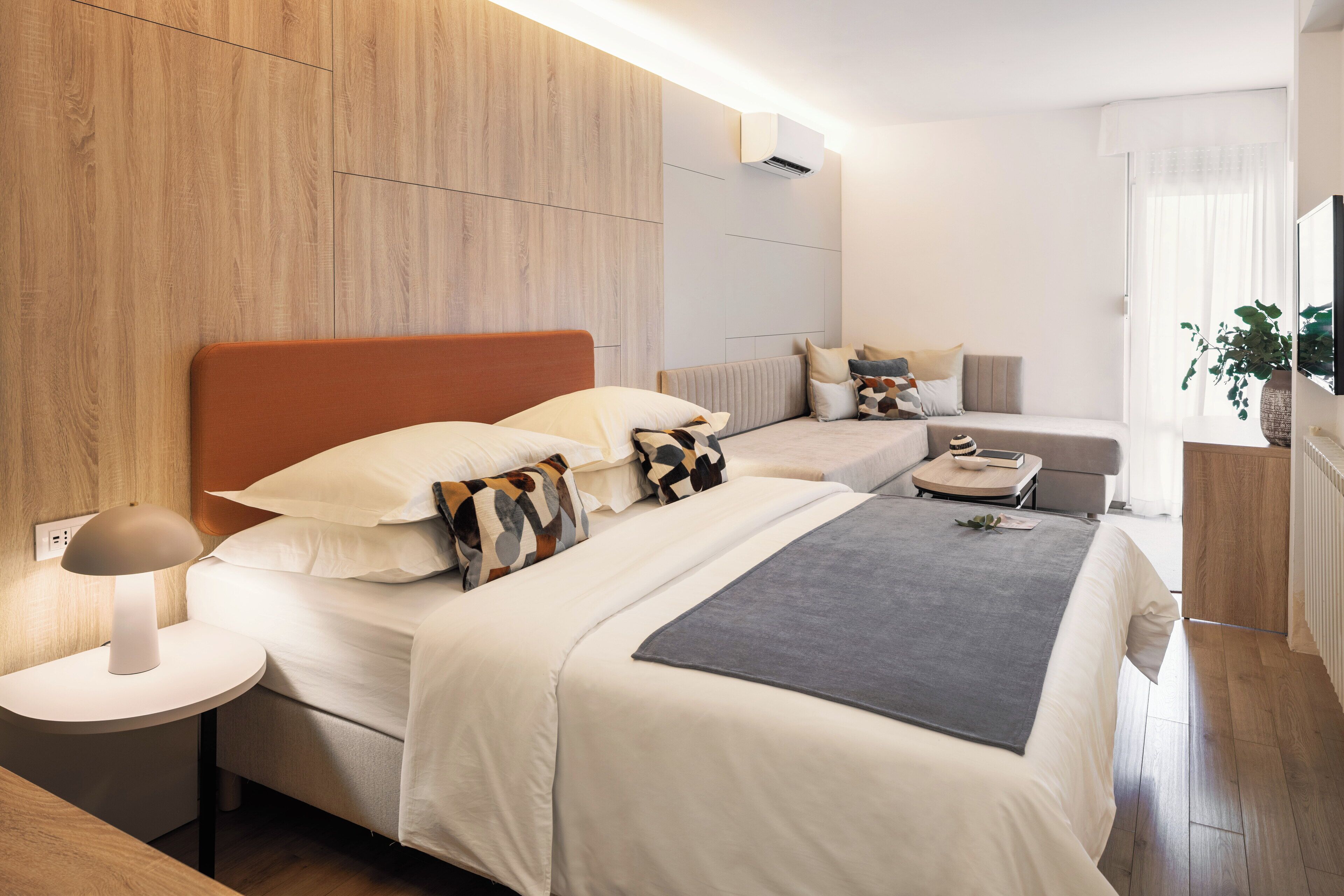 A modern hotel room with a large bed, a smaller bed, a bedside table with a lamp, and a view of the outdoors.