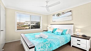 Un dormitorio con cama, mesitas de noche, ventilador de techo, ventana con persianas y un cuadro con temática playera en la pared.