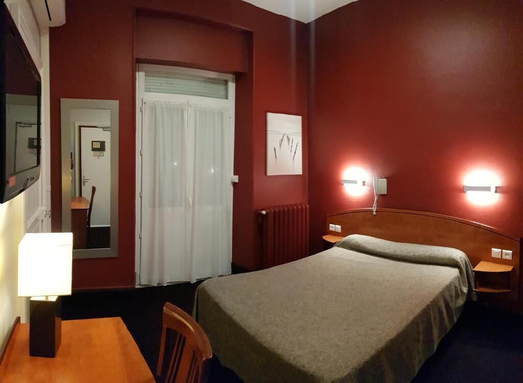 A hotel room with a bed, a desk, a chair, a mirror, a door, and a wall-mounted light fixture.
