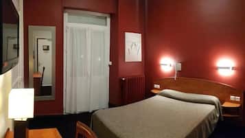 A hotel room with a bed, a desk, a chair, a mirror, a door, and a wall-mounted light fixture.