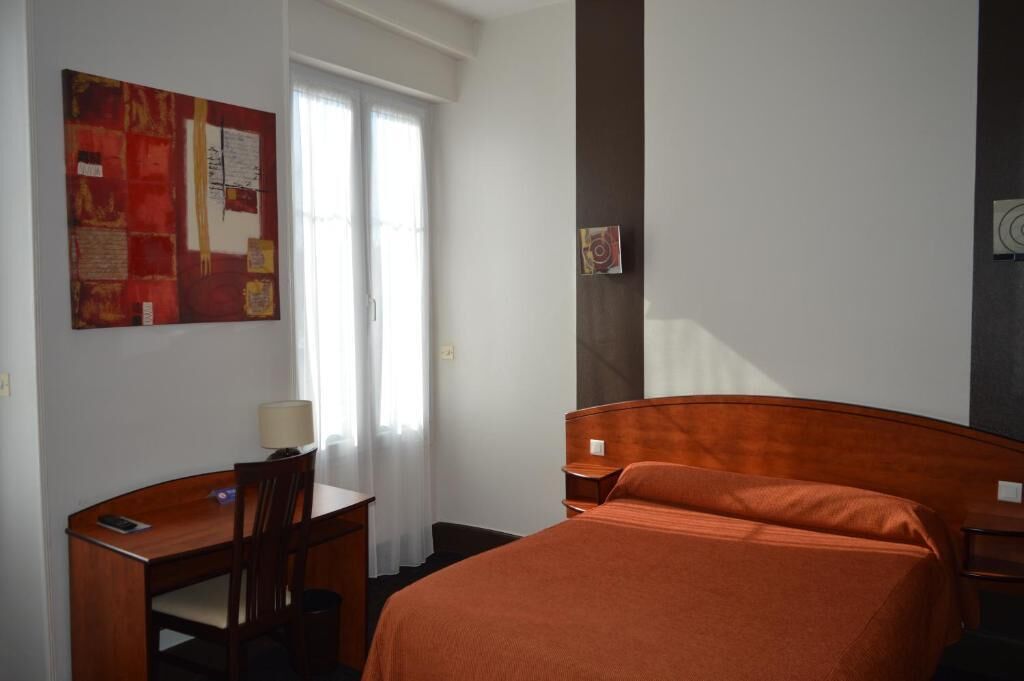 A hotel room with a bed, a desk, and a chair. There is a window with curtains, a wall with artwork, and a door.