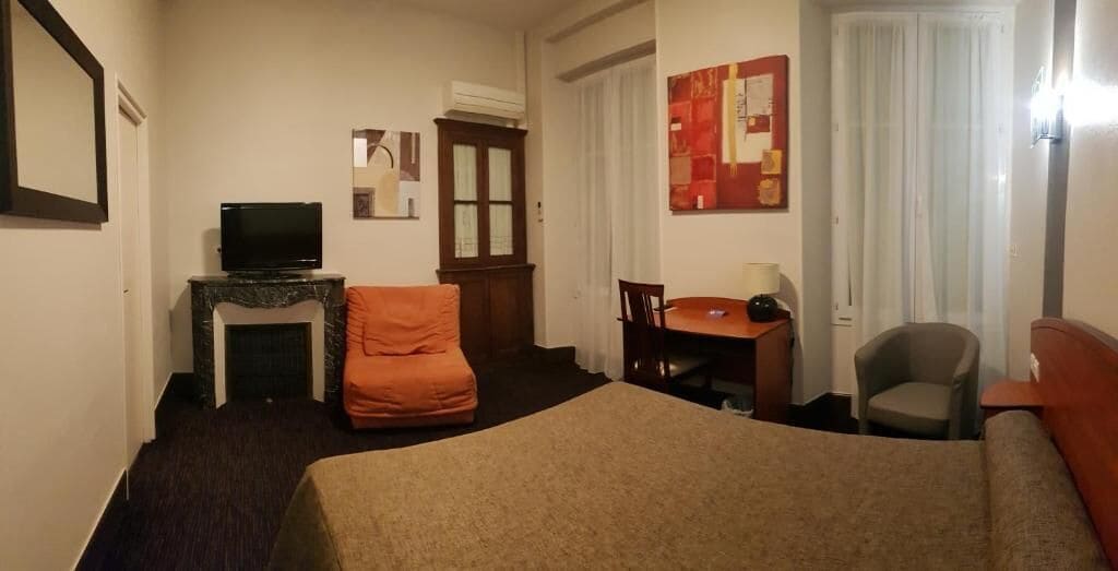 A hotel room with a bed, a television, a desk with a chair, a small table, and a window with curtains.