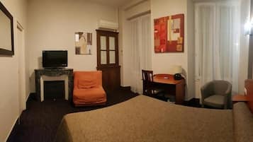 A hotel room with a bed, a television, a desk with a chair, a small table, and a window with curtains.