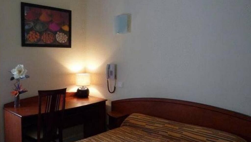 Una habitación de hotel con cama, un escritorio con lámpara, teléfono y un cuadro en la pared.
