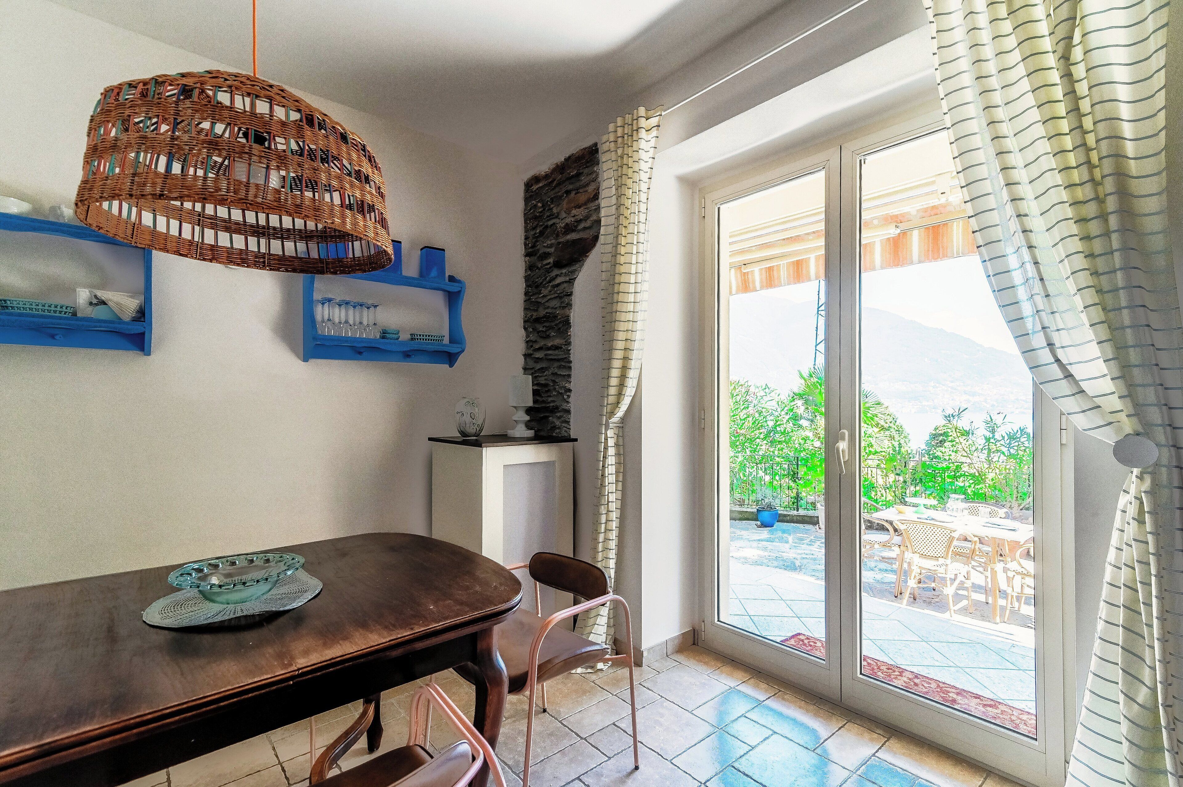 Un coin repas comprenant une table, des chaises, un luminaire suspendu en osier et une vue sur une terrasse extérieure, accessible par une porte vitrée.