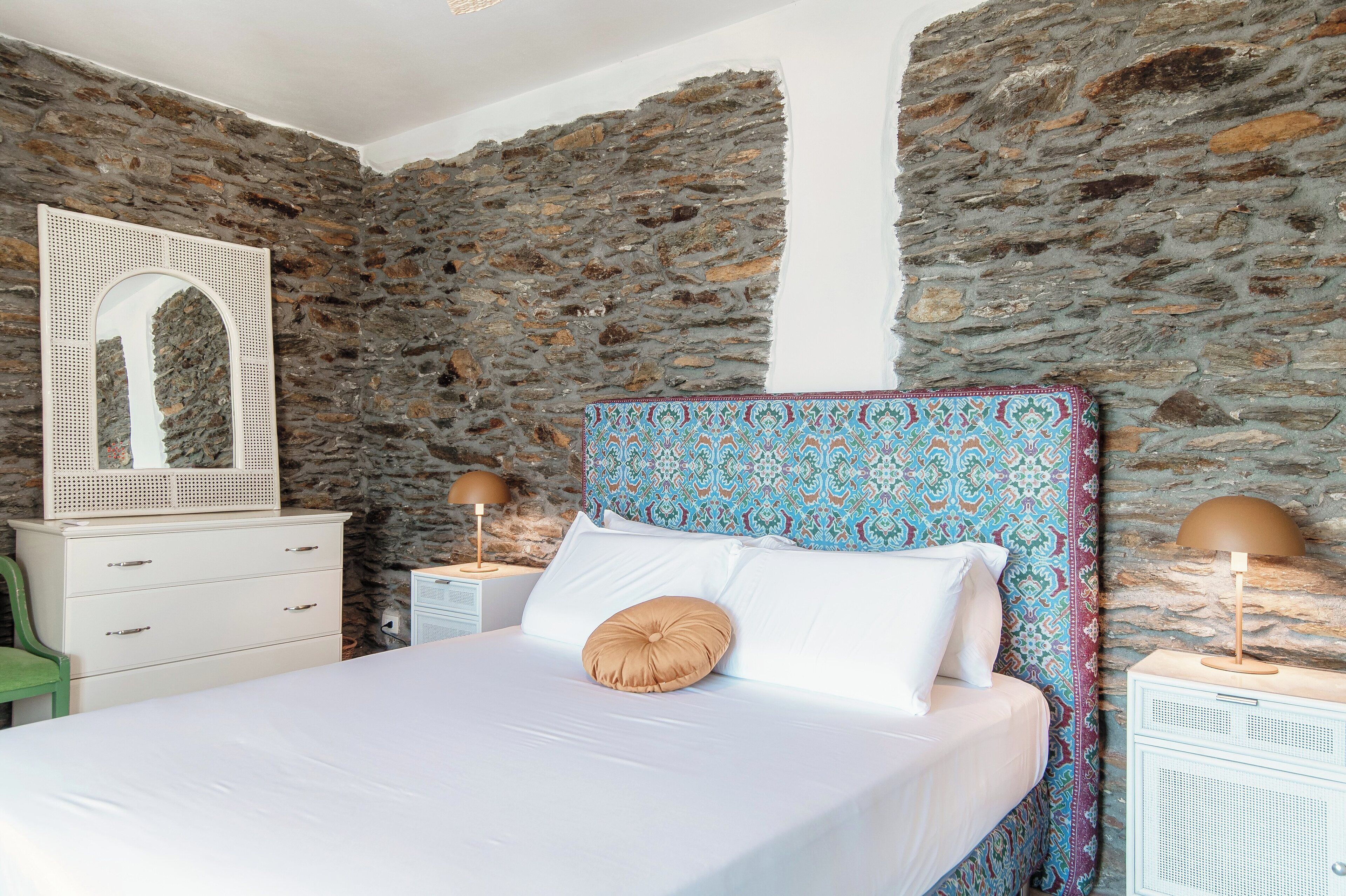 Een slaapkamer met een stenen muur, een bed met witte beddengoed, een gestreept hoofdeinde en twee bedlampen.