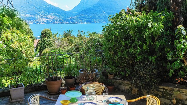 An outdoor dining area with a table set for two, surrounded by potted plants and overlooking a scenic lake and mountains. - Lost in Paradise  (San Siro)