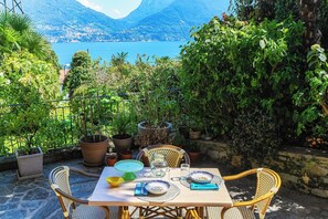 An outdoor dining area with a table set for two, surrounded by potted plants and overlooking a scenic lake and mountains. - Lost in Paradise  (San Siro)