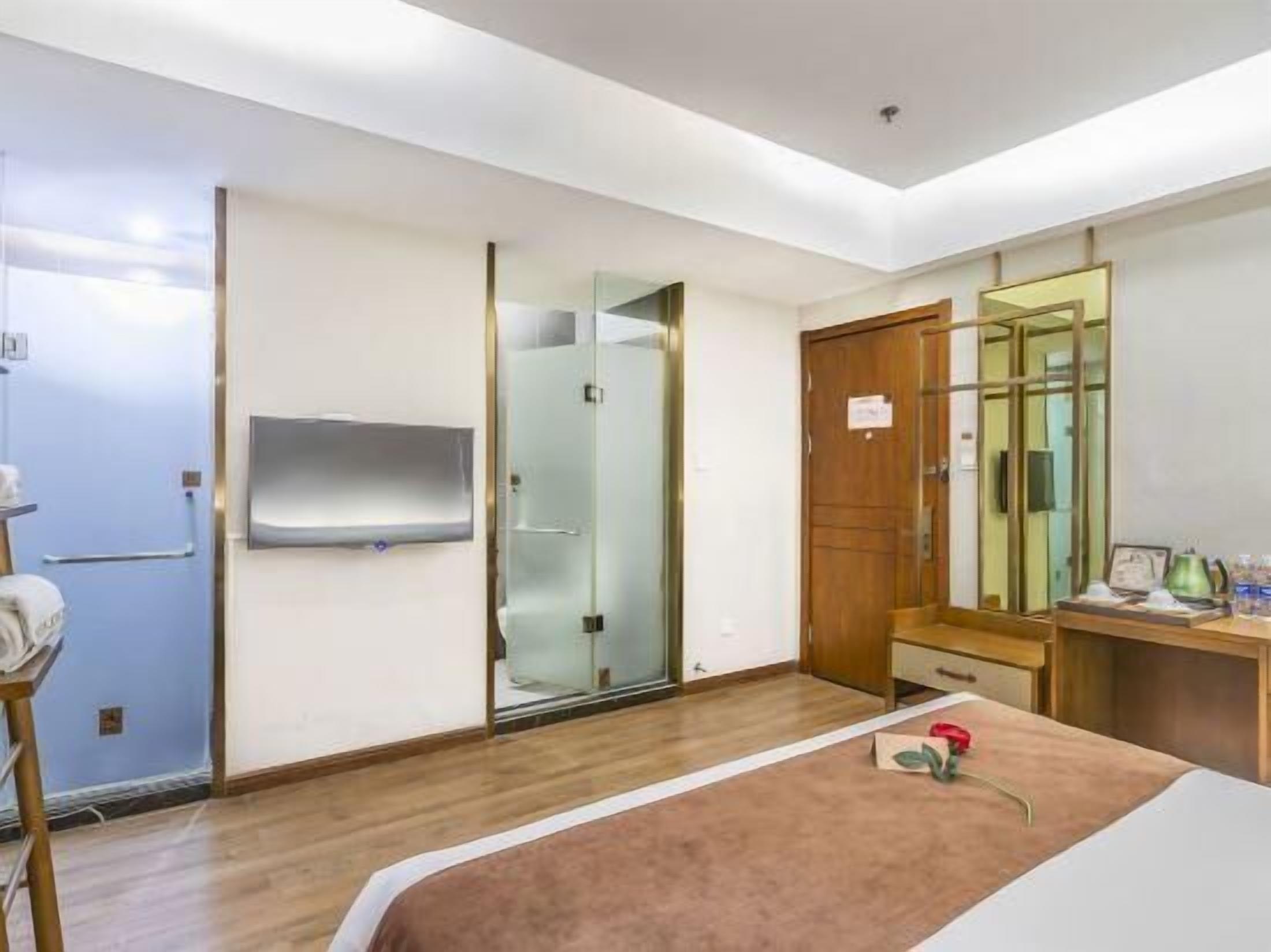 A hotel room with a wooden floor, a bed with a brown cover, a wooden desk with a mirror, a television mounted on the wall, and a sliding door leading to a bathroom.