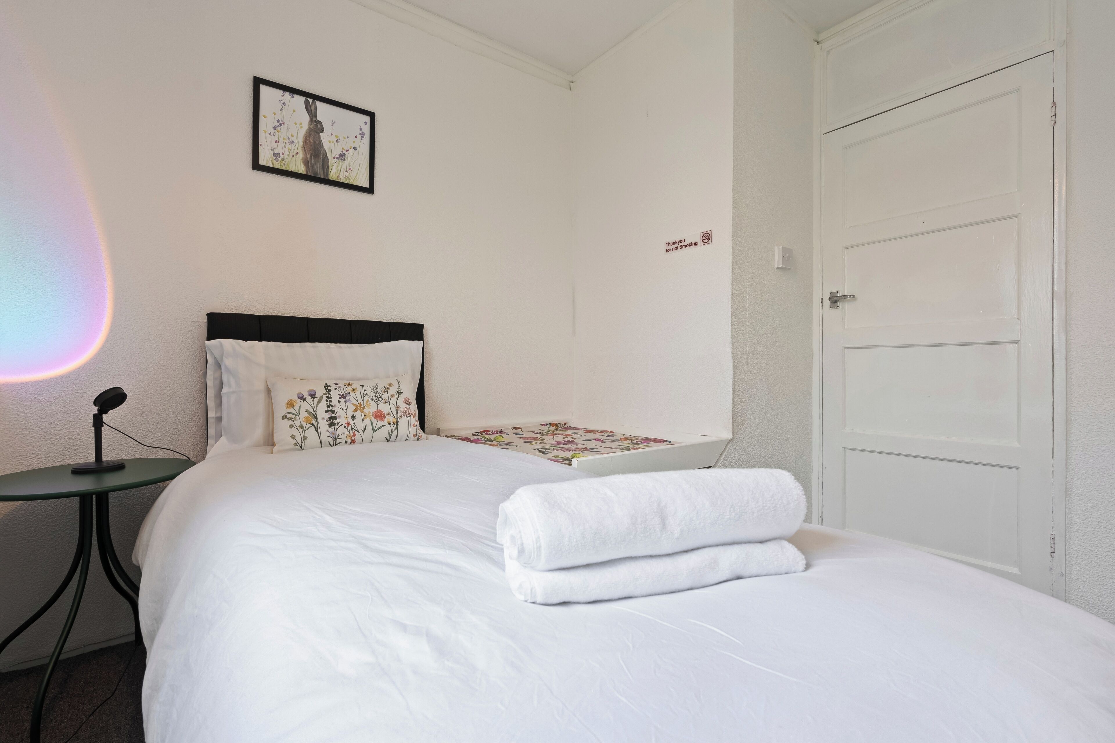 A neatly made bed with white linens and a folded towel, a small table with a microphone, a framed picture on the wall, and a closed door.
