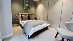 A modern bedroom with a large bed, a chair, a wardrobe, and a wall-mounted artwork.