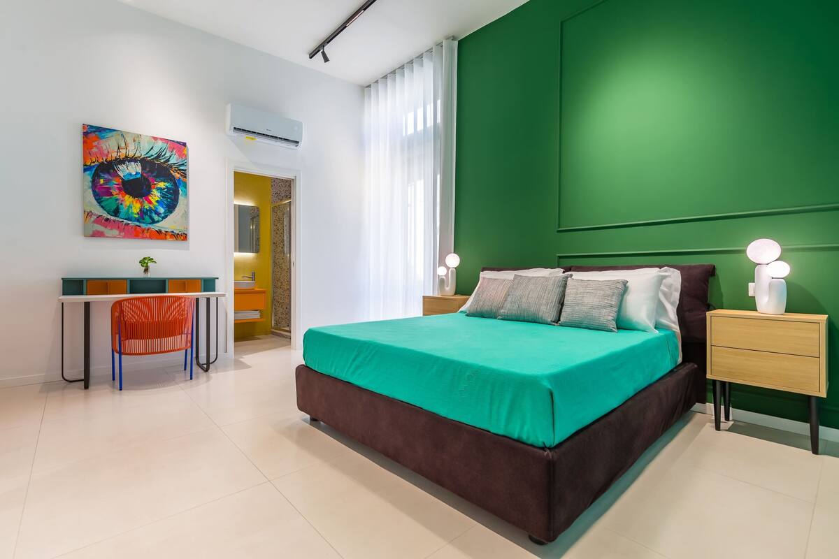 A modern bedroom with a large bed, a desk with a chair, a green accent wall, and a colorful artwork.