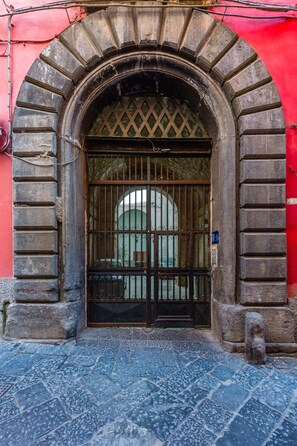 Un arc en pierre surmonté d’une grille métallique, menant à un bâtiment dont le mur est rouge.