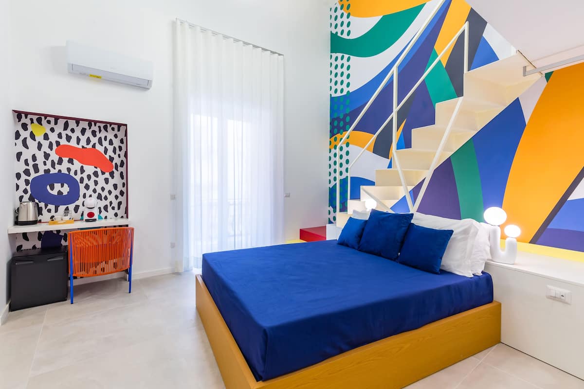 A modern bedroom with a large bed, a desk with a chair, a colorful abstract wall, and a staircase.