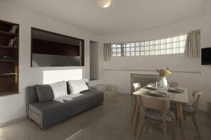 Ein modernes Wohnzimmer mit einem grauen Sofa, einem Esstisch mit Stühlen, einem Bücherregal und einem großen Fenster mit Vorhängen.