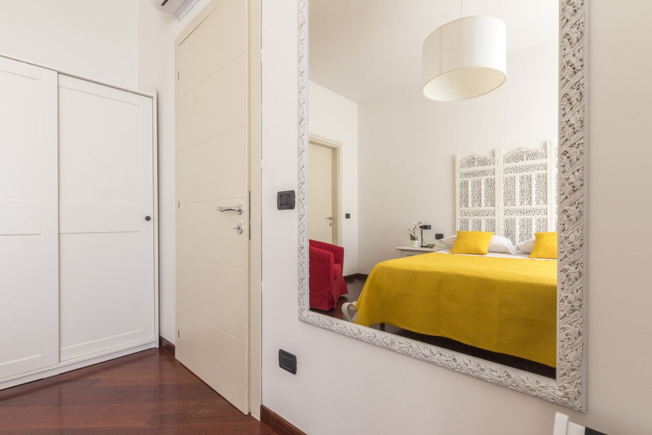Une chambre d’hôtel avec un grand miroir, un lit recouvert d’une couverture jaune, une serviette rouge et une porte blanche.